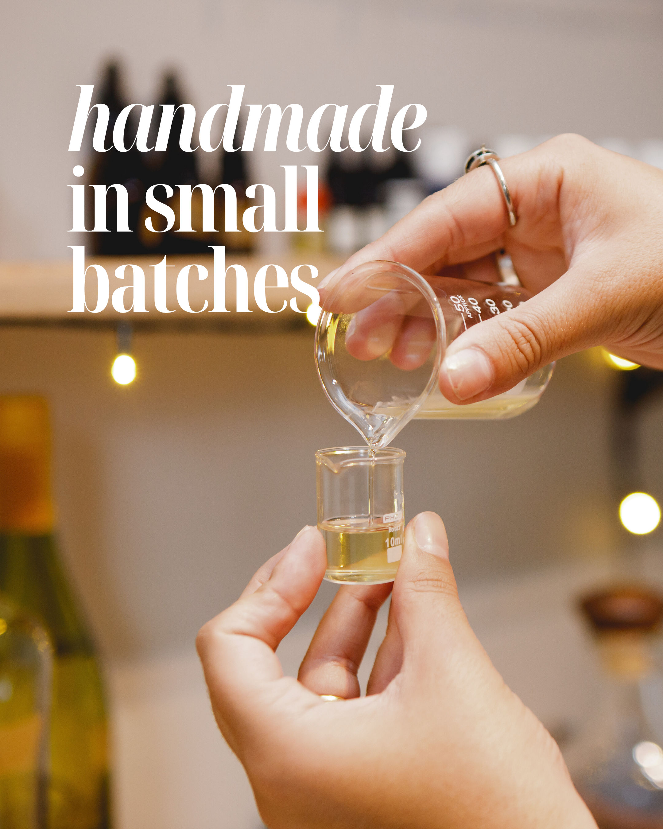 Hand pouring liquid from a small container into another, with text 'handmade in small batches' on a blurred background.